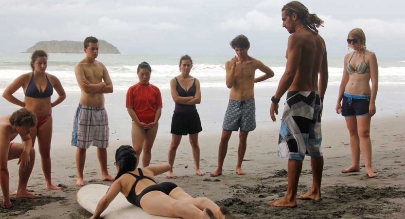 A group of students watch someone give a surf lesson on the sand. 